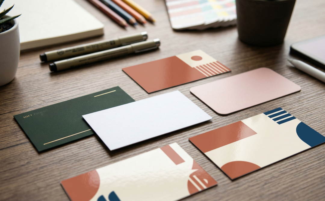 Un gros plan esthétique sur différentes cartes de visite de tailles standards (85x54mm, 89x51mm) avec des finitions variées (mat, brillant, soft touch) disposées sur un bureau de designer avec des outils de création.